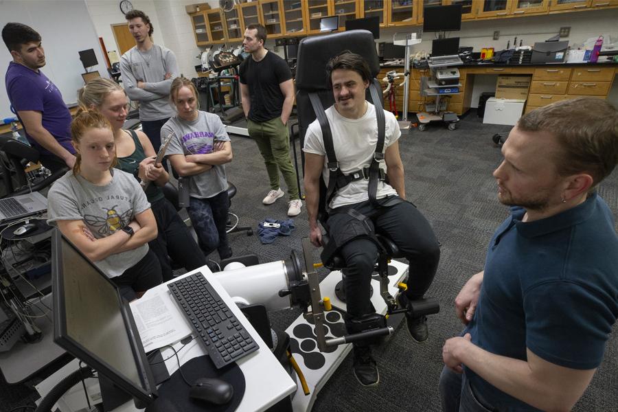 A faculty member and students gather around a student strapped in a chair to collect data during an exercise during class.